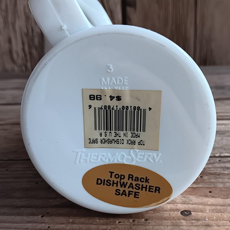 May include: A white plastic Thermo-Serv mug with a handle. The bottom of the mug has a sticker that reads "Top Rack Dishwasher Safe" and "Made in the U.S.A."