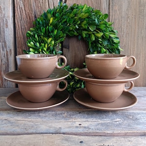 May include: Four brown ceramic teacups and saucers are stacked in pairs. The cups have a rounded shape with a handle. The saucers are round and flat. The background features a green wreath and a wooden surface.