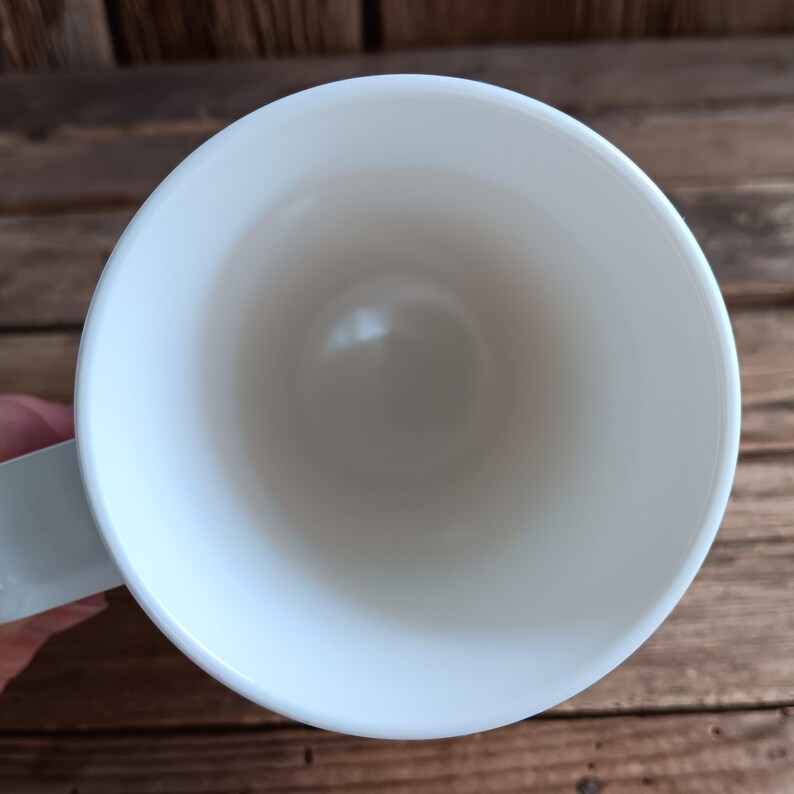 May include: A white ceramic mug with a pale blue handle, seen from above. The inside of the mug is smooth and white. The mug is set against a wooden background.