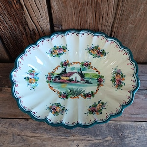 May include: Oval ceramic serving dish with a scalloped edge and a hand-painted design. The dish features a central landscape scene with a house and floral accents in pink, yellow, and green. The rim is teal, and the background is cream-colored.
