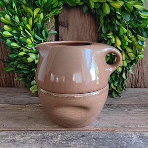 May include: A two-piece, brown ceramic gravy boat and warmer set. The gravy boat has a spout and handle, and sits atop a matching bowl with a curved indentation. The set is displayed against a backdrop of greenery and wood.
