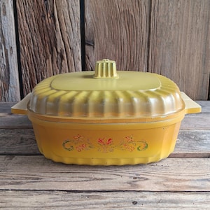 May include: A yellow vintage casserole dish with a matching lid. The rectangular dish has rounded edges and a floral pattern. The lid features a handle and a ribbed design. The dish is made of glass and is on a wooden surface.