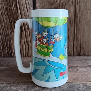 May include: A clear, tall beer mug with a white handle and rim. The mug features a colourful illustration of Goofy and Donald Duck in a hot air balloon, with a castle in the background. The base of the mug has a blue and white floral design, with the text "Walt Disney World".