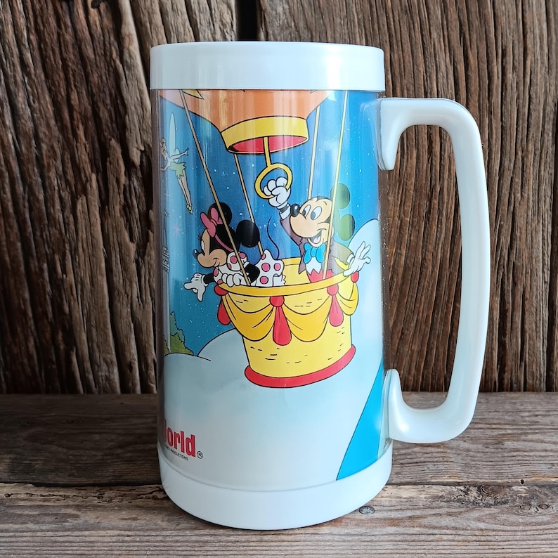 May include: A large mug with a white handle, depicting Mickey and Minnie Mouse in a hot air balloon. The balloon is yellow and red, set against a blue sky with clouds. The mug has a white lid and a light blue base.