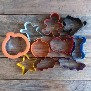 Puede incluir: Un conjunto de diez cortadores de galletas de metal en varias formas y colores, incluyendo un fantasma, una calabaza, un hombre de jengibre, un hueso y una estrella. Los cortadores están dispuestos sobre una superficie de madera.