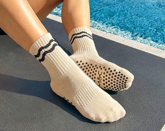 May include: A pair of beige yoga socks with black stripes and black grippy dots on the bottom. The socks are worn by a person sitting by a pool.