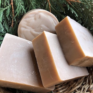 May include: A collection of handmade soaps in a woven basket, with a sprig of green foliage. The soaps are various shapes, including rectangular bars and a round soap. The soaps are a light brown color.