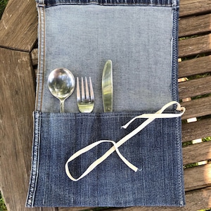 May include: A denim cutlery holder with a spoon, fork, and knife inside. The holder is made from blue jean material and has a white ribbon tie. The background is a wooden table.