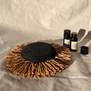 Puede incluir: Un difusor de piedra de lava negra con una base de paja trenzada. Dos pequeños frascos de vidrio ámbar de aceite esencial están al lado del difusor. Los frascos están etiquetados con "Huile essentielle de Vetiver naturelle 5 ml".
