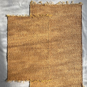 Puede incluir: Dos esteras rectangulares tejidas en tonos marrones y beige. Las esteras tienen una superficie texturizada y están rematadas con hilos amarillos a lo largo del borde superior. Las esteras son de diferentes tamaños, una se superpone parcialmente a la otra.