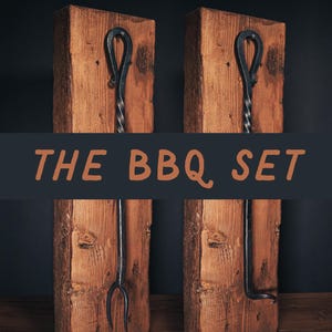 May include: The BBQ Set. Two sets of grilling tools are mounted on rustic wooden blocks. Each set includes a poker, a fork, and a spatula. The tools are made of dark metal with twisted handles and are displayed against a dark background.