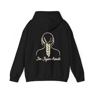 May include: Black hoodie featuring a cream-colored illustration of a braid and the back of a head. The text "Jin Jiyan Azadi" is printed below the illustration in a cursive font. The hoodie has a hood and long sleeves.