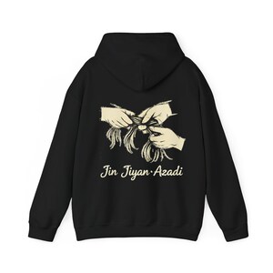 May include: Black hoodie with a cream-colored graphic on the back. The graphic depicts two hands braiding hair, with the words "Jin Jiyan Azadi" in a cursive font below the image. The hoodie has a hood and long sleeves.