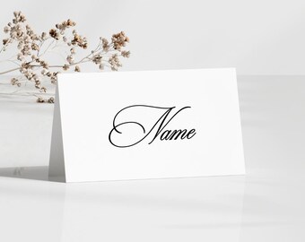 May include: A white place card with the word "Name" in elegant black script. The card is angled, suggesting it's standing on a surface. Dried floral accents are visible in the background, adding a touch of natural decor.