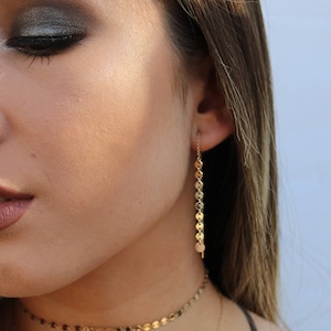 May include: Gold-toned, dangling earrings with a chain of small, circular charms. The earrings are shown with a matching choker necklace. The image is a close-up, highlighting the jewelry's delicate design and the warm tones of the metal.