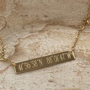 May include: A gold necklace with a bar pendant engraved with the coordinates "41° 56' 58" N 88° 01' 47" W".