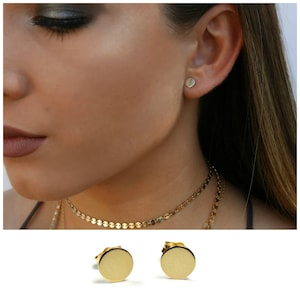 May include: A pair of gold-toned stud earrings with a round, flat design. The earrings are shown on a model's ear.