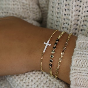 May include: Three gold bracelets on a wrist. The top bracelet features a small, diamond-encrusted cross charm. The middle bracelet has a chain with small, round, gold links. The bottom bracelet has a delicate, chain-link design.