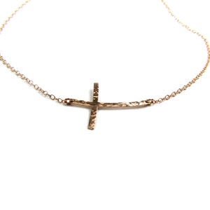 May include: A delicate gold necklace with a hammered cross pendant. The cross is sideways and has a slightly curved shape.