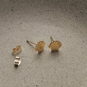 May include: Gold seashell stud earrings with matching earring backs. The earrings are made of 14K solid gold. The image also includes the text "Gems in Vogue Handmade Jewelry".