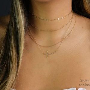 May include: A layered gold necklace set. The top layer is a delicate chain with small, flat discs. The second layer is a simple chain. The third layer features a curved bar pendant with small, sparkling stones. The model is wearing a white top.