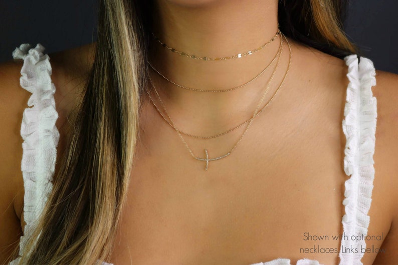 May include: A layered gold necklace set. The top layer is a delicate chain with small, flat discs. The second layer is a simple chain. The third layer features a curved bar pendant with small, sparkling stones. The model is wearing a white top.