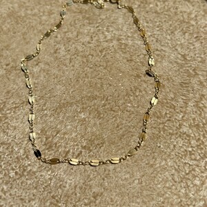May include: A delicate gold-colored necklace with a chain of small, flat, oval-shaped links. The necklace is displayed on a textured, light brown surface. The clasp is visible at the top of the necklace.