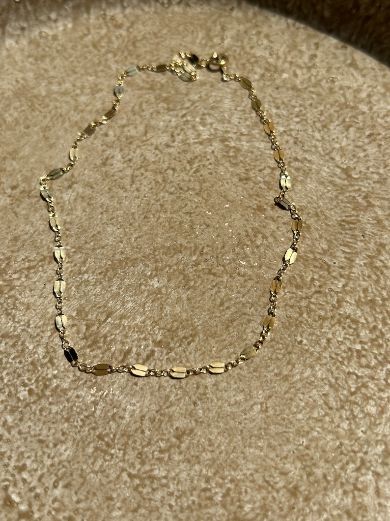 May include: A delicate gold-colored necklace with a chain of small, flat, oval-shaped links. The necklace is displayed on a textured, light brown surface. The clasp is visible at the top of the necklace.