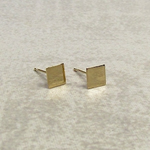May include: A pair of small, square gold stud earrings. Each earring features a flat, square design with a brushed texture. The posts are thin and extend from the back of the square. The earrings are displayed on a light-colored surface.