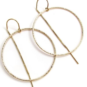 May include: A pair of gold-colored hoop earrings with a textured surface. Each earring features a large, open circle design with a straight bar and a chain-like element. The earrings are displayed against a white background.