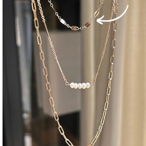 May include: A layered necklace set featuring three strands of varying designs. The top strand has small, flat, oval-shaped links. The middle strand has a row of white pearls. The bottom strand has large rectangular links. The necklaces are gold-colored.
