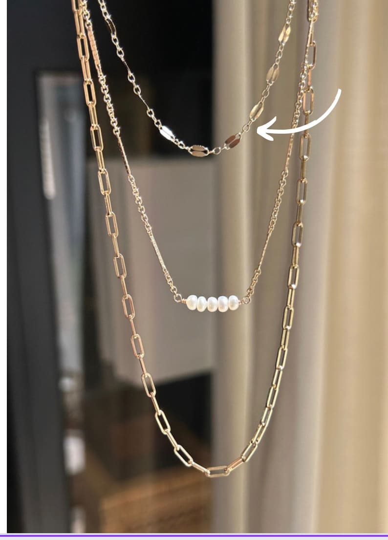 May include: A layered necklace set featuring three strands of varying designs. The top strand has small, flat, oval-shaped links. The middle strand has a row of white pearls. The bottom strand has large rectangular links. The necklaces are gold-colored.