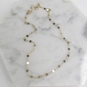 May include: Delicate gold-colored necklace with a repeating oval and rectangular link design. The necklace is displayed on a white marble surface, showcasing its elegant and minimalist style. The chain is approximately 16 inches long.