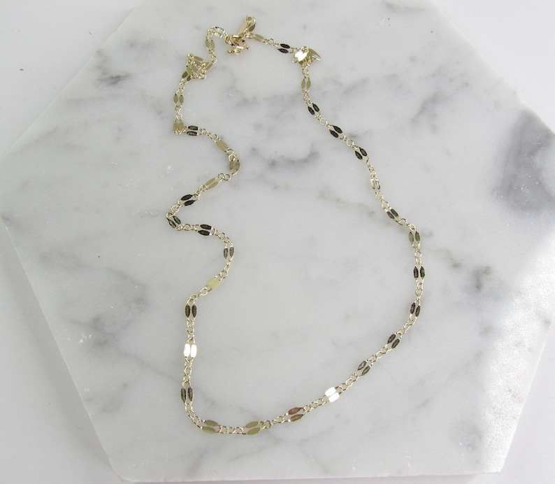 May include: Delicate gold-colored necklace with a repeating oval and rectangular link design. The necklace is displayed on a white marble surface, showcasing its elegant and minimalist style. The chain is approximately 16 inches long.