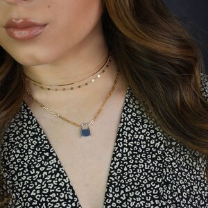 May include: A gold-toned necklace set featuring three layers. The top layer is a delicate chain, the middle layer has small rectangular accents, and the bottom layer has a padlock pendant with a dark blue center. The model is wearing a black and white patterned top.
