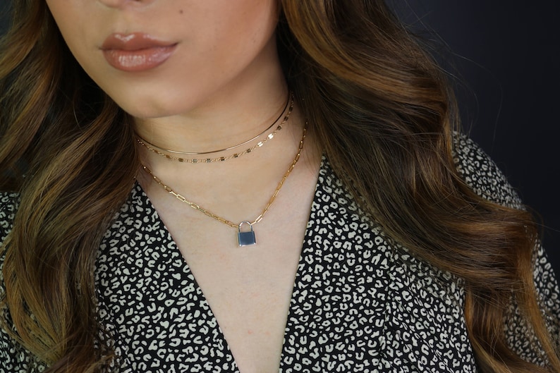May include: A gold-toned necklace set featuring three layers. The top layer is a delicate chain, the middle layer has small rectangular accents, and the bottom layer has a padlock pendant with a dark blue center. The model is wearing a black and white patterned top.
