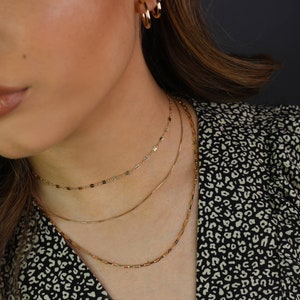 May include: Three gold necklaces of varying designs are displayed. The top necklace is a delicate chain with small, rectangular links. The middle necklace is a simple, thin chain. The bottom necklace features small, rectangular bars. Gold hoop earrings are also visible.