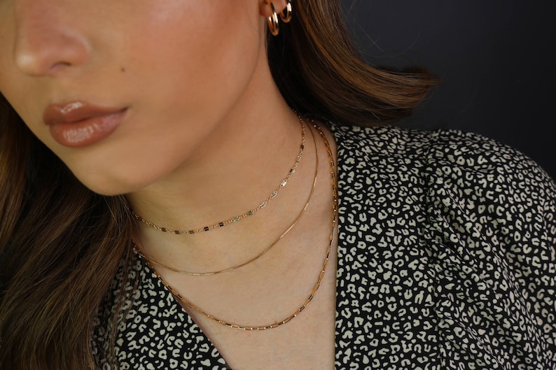 May include: Three gold necklaces of varying designs are displayed. The top necklace is a delicate chain with small, rectangular links. The middle necklace is a simple, thin chain. The bottom necklace features small, rectangular bars. Gold hoop earrings are also visible.