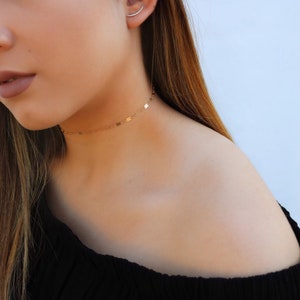 May include: A delicate gold choker necklace with small, rectangular charms. The necklace is worn around the neck and sits just above the collarbone. The jewelry is simple and elegant, suitable for everyday wear or special occasions.
