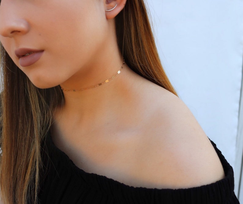 May include: A delicate gold choker necklace with small, rectangular charms. The necklace is worn around the neck and sits just above the collarbone. The jewelry is simple and elegant, suitable for everyday wear or special occasions.