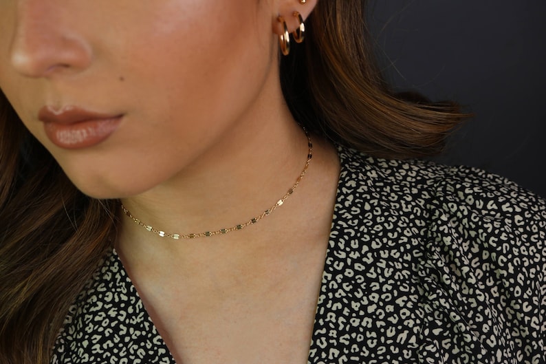 May include: A gold-colored choker necklace with small, rectangular links. The necklace is worn on a person with gold hoop earrings and a black and white patterned top. The background is a dark gray.