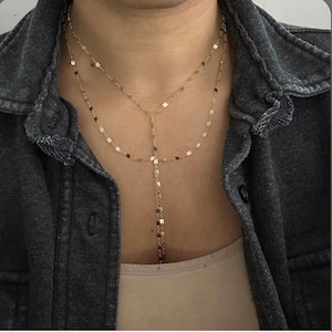 May include: A gold layered necklace with a long, thin chain and a shorter, thicker chain. The necklace is made of small, flat, oval-shaped links.