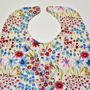 May include: A white bib with a watercolor floral pattern. The bib features a variety of colorful flowers, including red, blue, and pink, with green stems and leaves. The bib has a curved neckline and a long, rectangular shape.