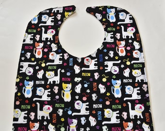 Puede incluir: Babero negro con un patrón de gato colorido. El babero tiene una abertura de cuello blanca y está cubierto de gatos con la palabra "MEOW" repetida en todas partes.