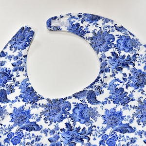 May include: A white bib with a blue floral pattern. The bib has a rounded neckline and is made of a soft, comfortable fabric.