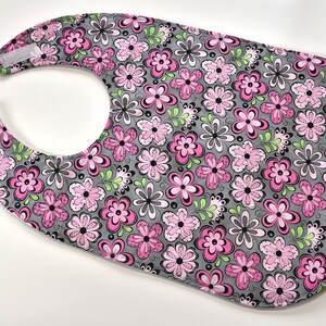 May include: A gray bib with a pink and green floral pattern. The bib has a hook and loop closure.