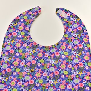 May include: A purple bib with a floral pattern featuring pink, green, yellow, and white flowers, and pink butterflies. The bib has a curved neckline and a Velcro closure.