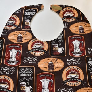 May include: A brown and orange baby bib with a coffee theme. The bib features a variety of coffee-related images and text, including coffee cups, coffee beans, coffee grinders, and the words "Coffee Time" and "House Blend".