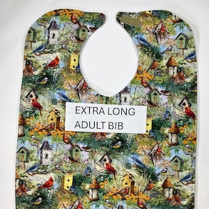 May include: An extra-long adult bib with a colorful bird and birdhouse pattern. The bib features a large neck opening and is made of a soft, absorbent fabric. The text "EXTRA LONG ADULT BIB" is printed on the front.