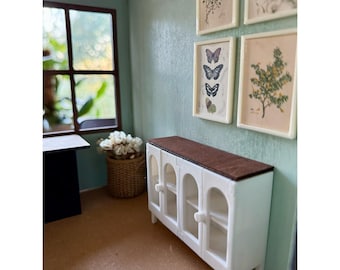 Puede incluir: Miniatura de armario blanco con puertas de cristal y tapa marrón. El armario está en una habitación de casa de muñecas con grabados botánicos enmarcados y una ventana. Una cesta tejida con flores blancas está cerca.
